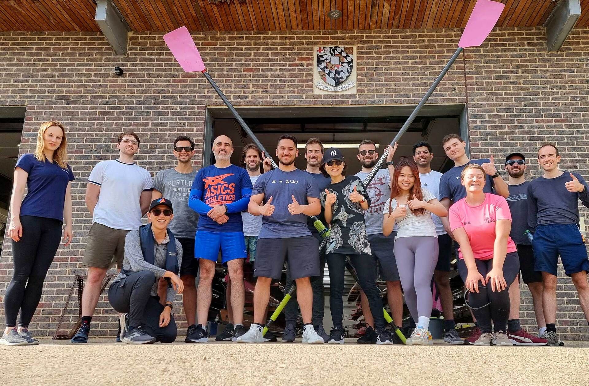Several people in workout gear smile in front of a boat house. Two support crossed oars with black shafts and red blades.