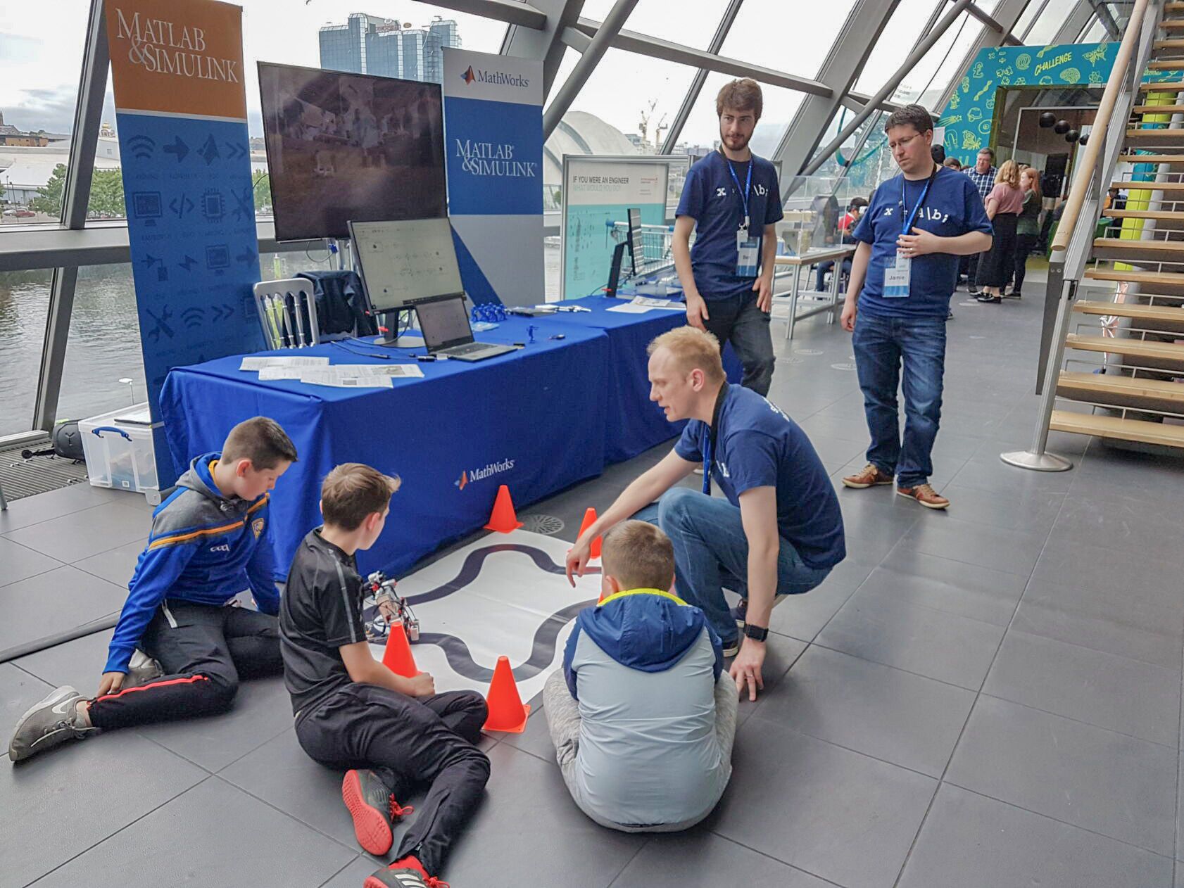 People gather around a small robot track at a MATLAB and Simulink booth in a modern building.