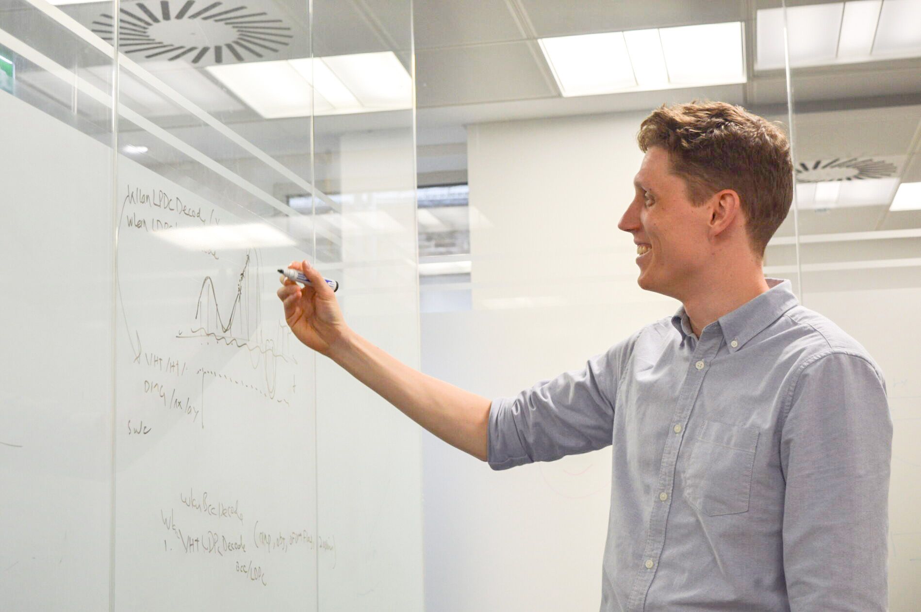 A person in a blue shirt writes on a glass board with a marker, smiling in a bright office setting.
