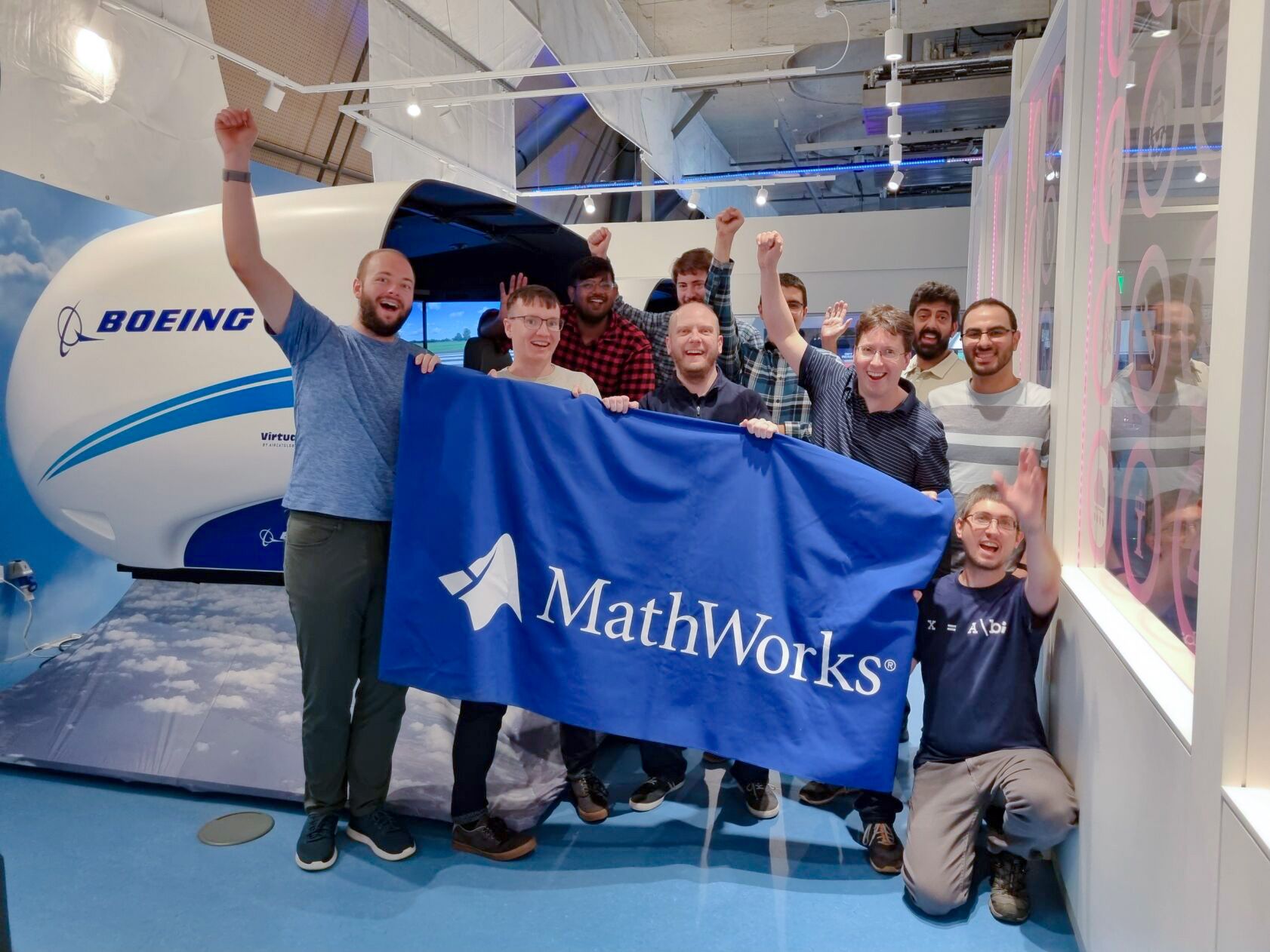 A group of people cheerfully hold a MathWorks banner in front of a Boeing simulator.