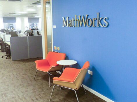 Two orange chairs  and a small white table sit in an office lobby with a blue wall featuring the MathWorks logo.