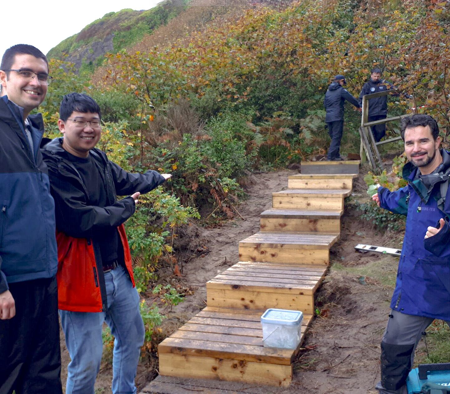 Three people smile and point at newly constructed wooden steps on a hillside, with two others working in the background.