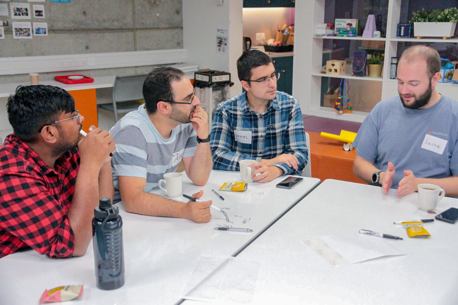 Four men sit at a table, engaged in discussion, with mugs and pens in front of them.