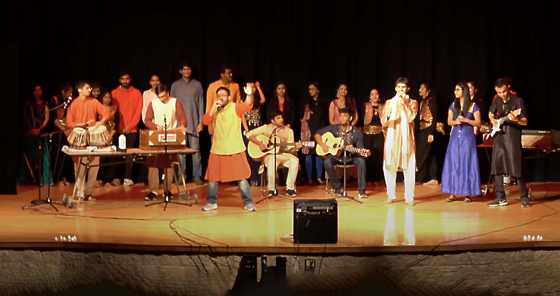 Group photo of Desi Utsav performers on stage with drums, guitars, and other instruments.