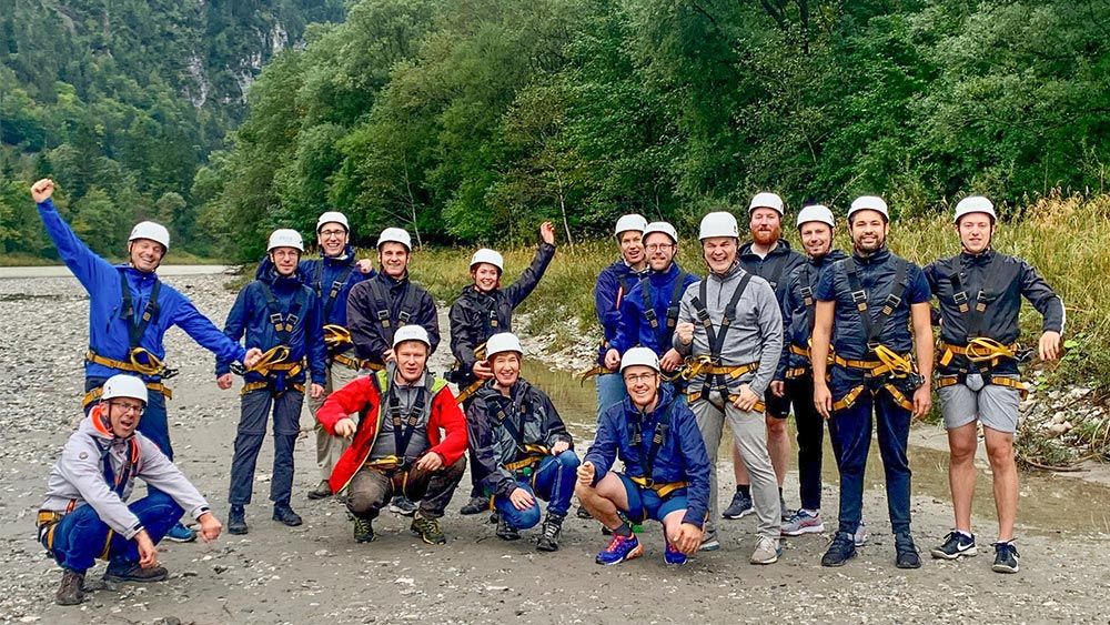 Group of 16 MathWorks staff wearing zipline harnesses and helmets, standing on a dirt road in a forest.